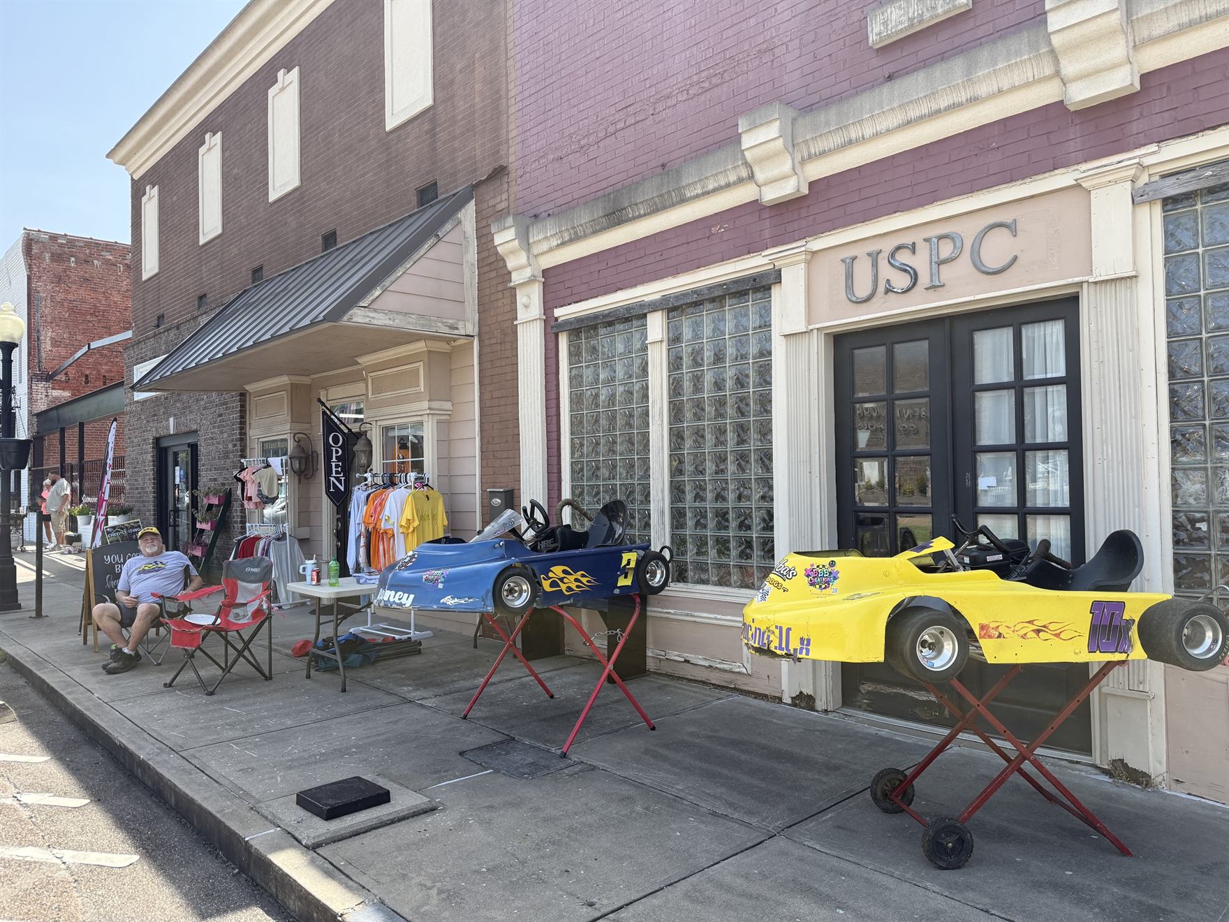 Ronnie Rainey Sr. sitting out front by the karts with the Open flag flying and merch racks along the storefront