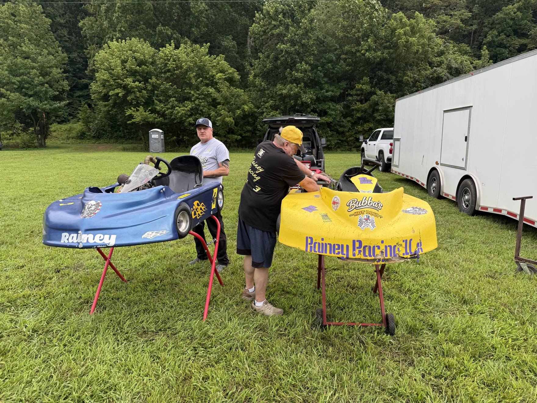Travis's yellow 10X and Ronnie's blue 3 karts up on stands in the grass pits at Sleepy Hollow Speedway, with the team working on them