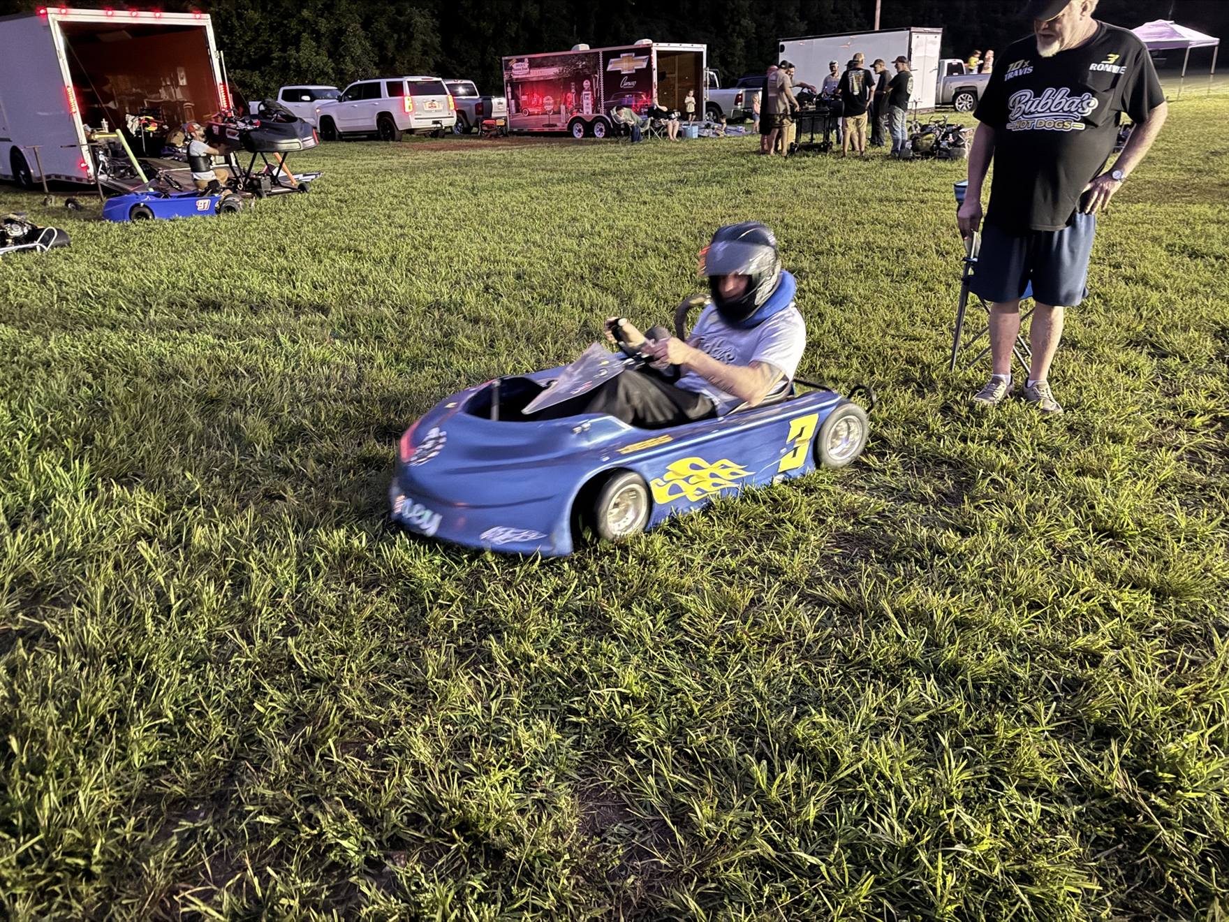 Ronnie fired up in the blue 3 kart in the pits at night, with Ronnie Sr. in a Bubba's Hot Dogs shirt looking on