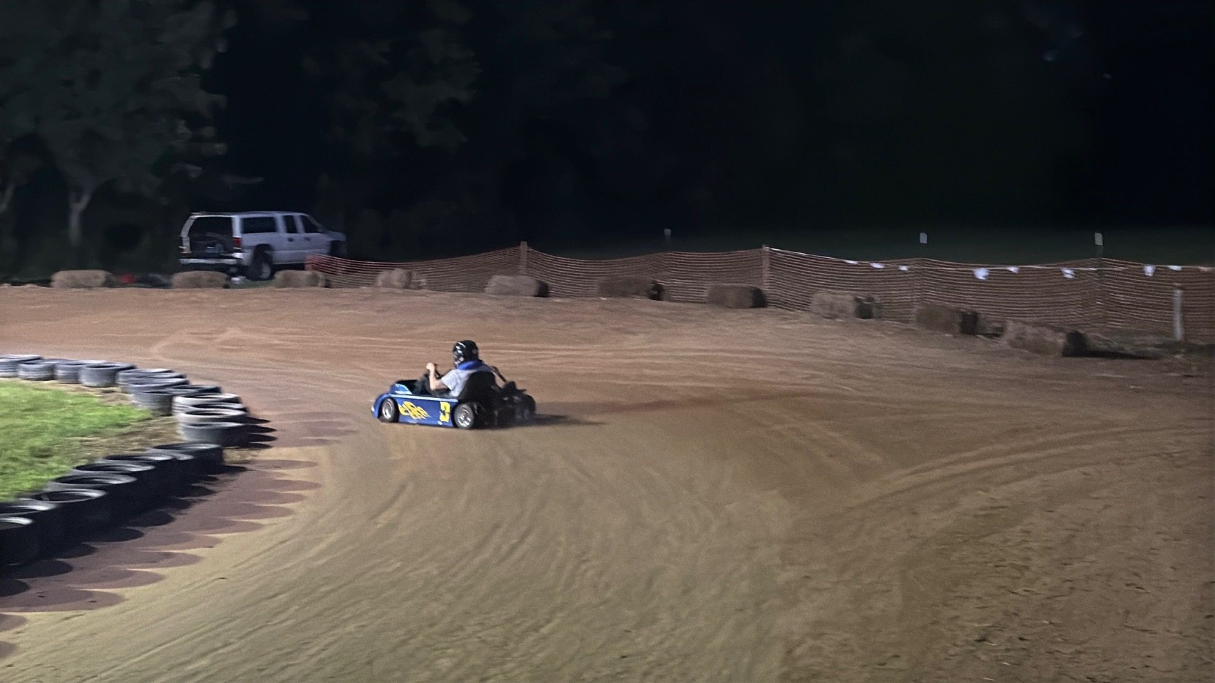 The blue 3 kart running the bottom of the dirt track at night past the tire wall