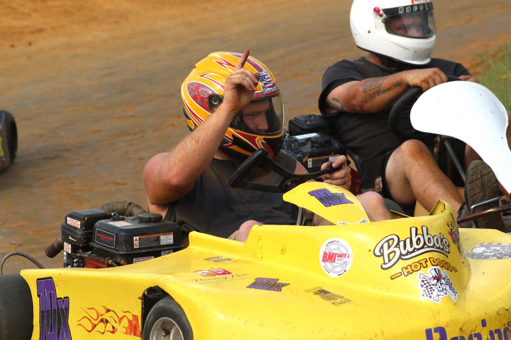 Travis pointing a finger to the sky from the seat of the 10X alongside another kart on track